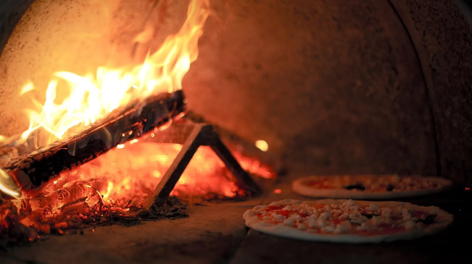 Brændefyret stenovn set indefra: flammer og gløder til venstre og to pizzaer, der bager på den murede ovnbund