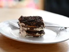 Et stykke tiramisu med lyse cremelag og mørkt kakaodrys på en hvid tallerken