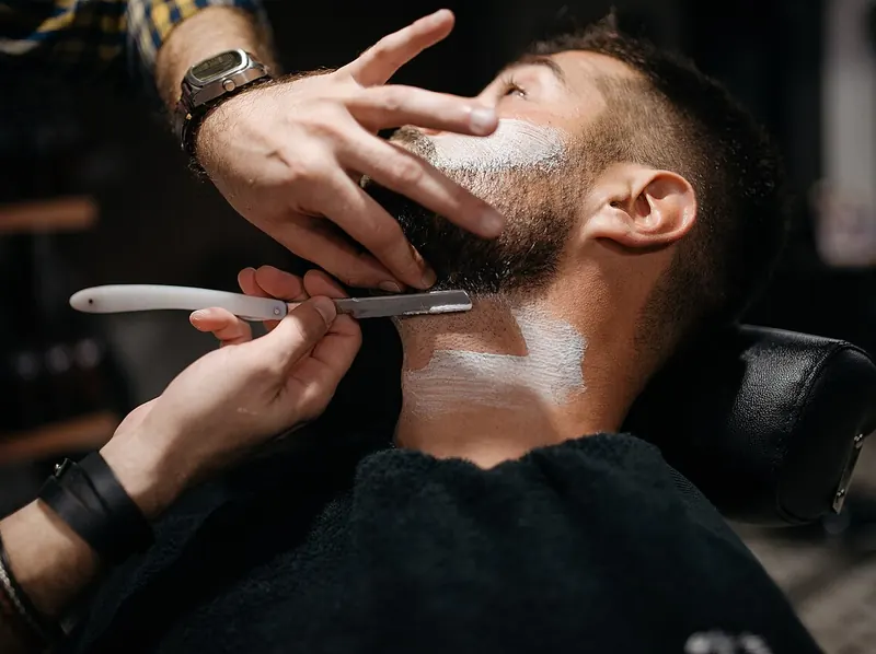 En mand ligger tilbagelænet i barberstolen med barberskum på hals og kinder, mens barberen fører en hvid barberkniv hen over halsen og holder hagen med den anden hånd