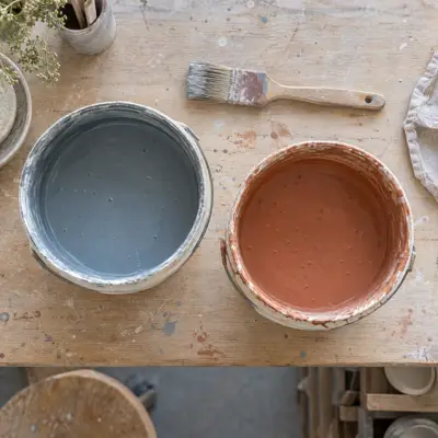 Åbne spande med flydende glasur i blågrå og terracotta med en pensel over, set lige oppefra